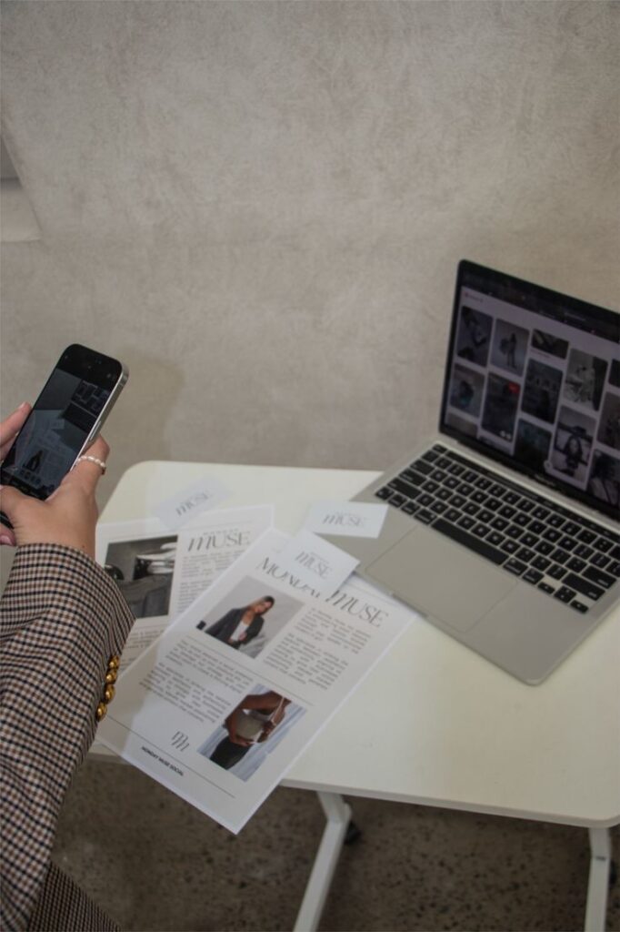 Person using smartphone near laptop and printed documents on a desk, focusing on fashion content research.