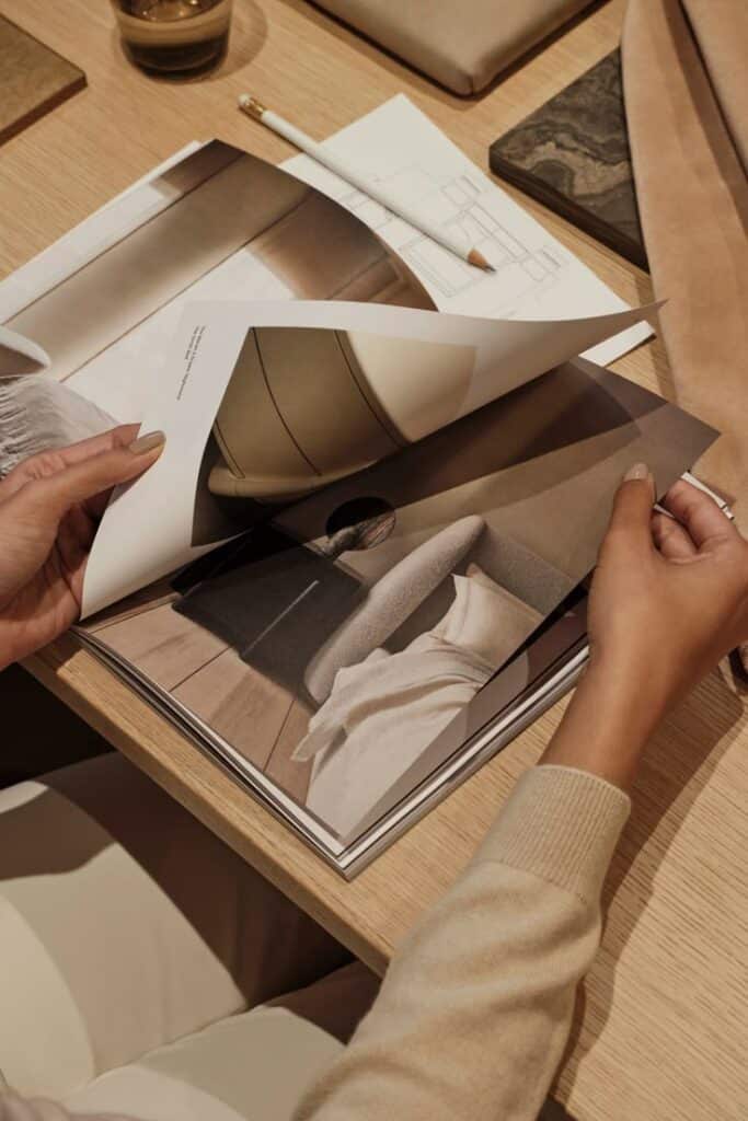 Person flipping through interior design magazine at a wooden desk, with sketches and materials around.