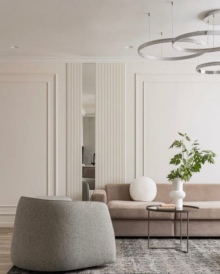 Modern living room with beige sofa, gray armchair, decorative plant, and contemporary lighting design.