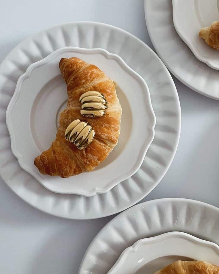 Golden croissant and shiny rings on white plates, stylish breakfast display.