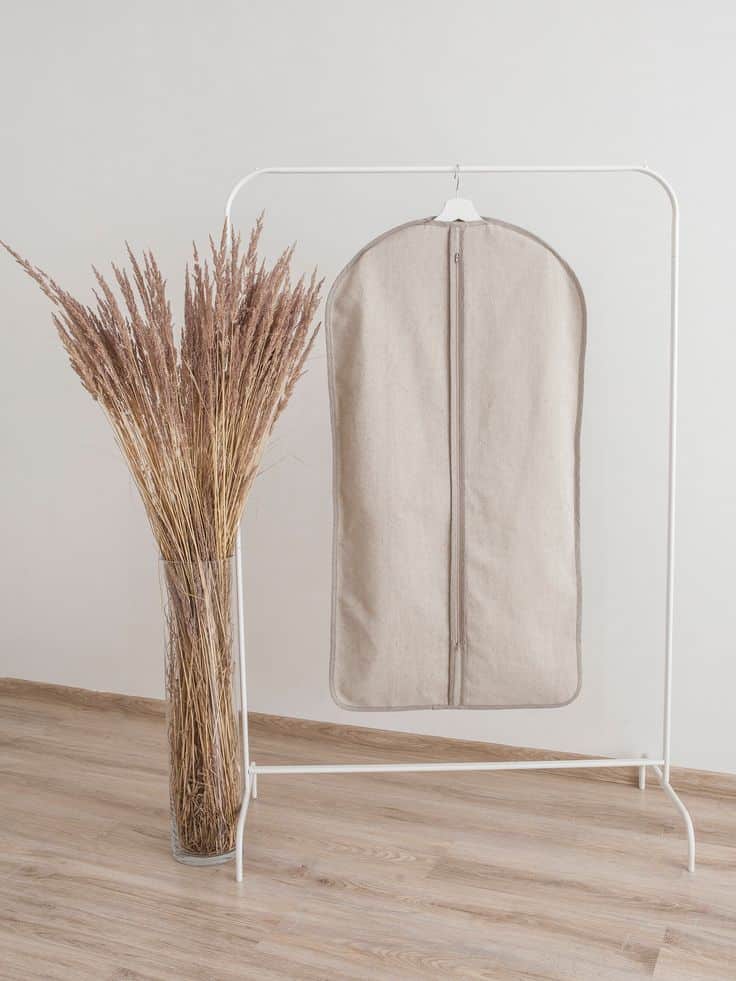 Beige garment bag on a hanger with tall dried grasses in a vase, minimalist home decor.