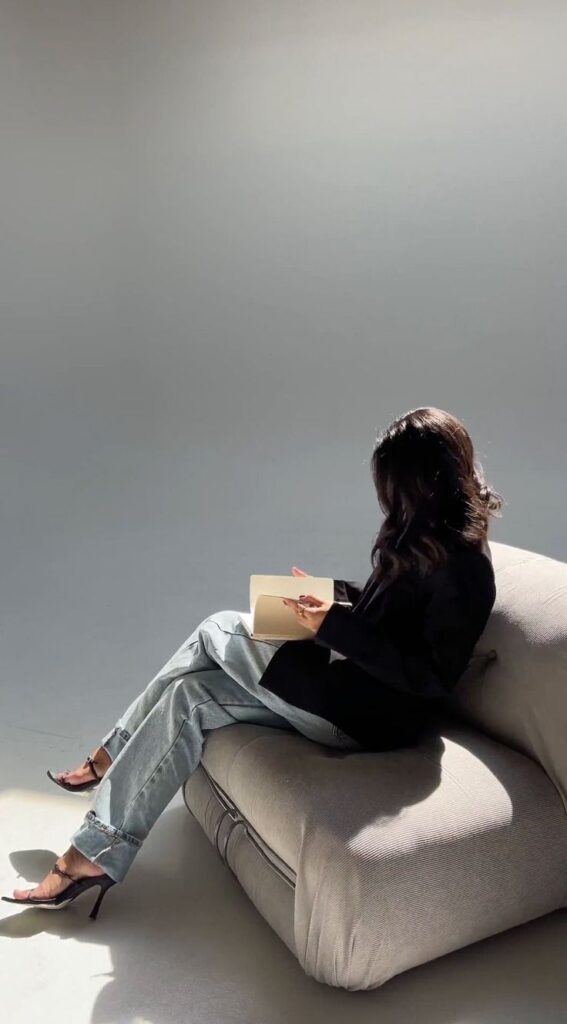Woman reading a book on a cushioned chair, wearing jeans and black top, in a minimalist setting.