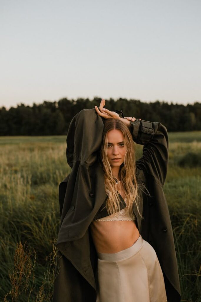 Woman in stylish coat posing in a serene field at sunset, blending fashion and nature.