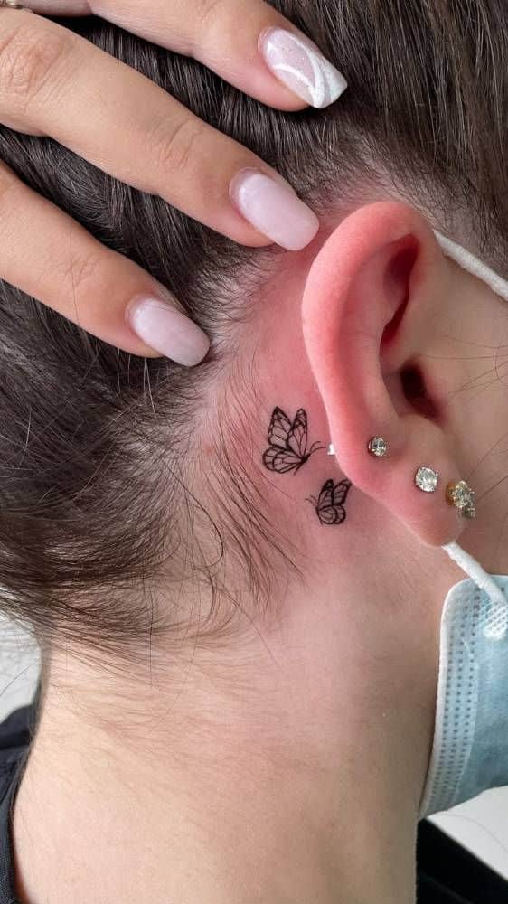 Small butterfly tattoo behind ear with elegant nail art and multiple ear piercings.