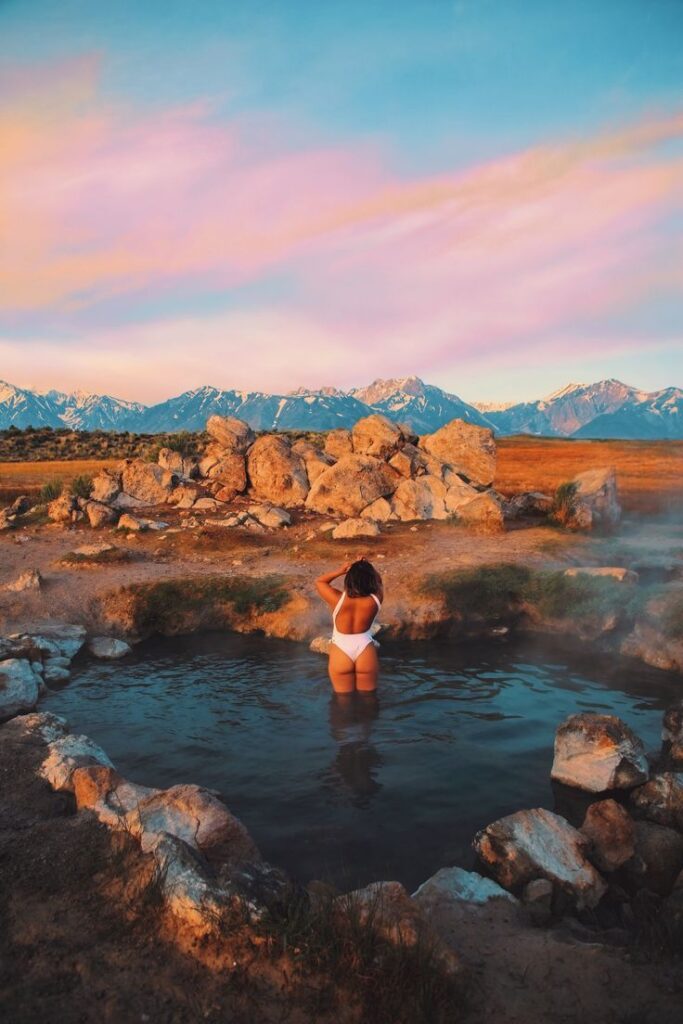 Woman enjoying a natural hot spring with mountains and colorful sky at sunset.