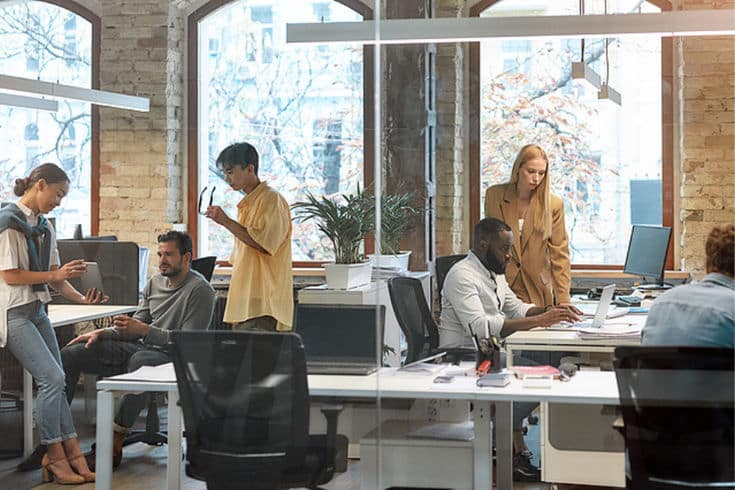 Open-plan office with diverse team collaborating, discussing, and working on laptops at modern desks.