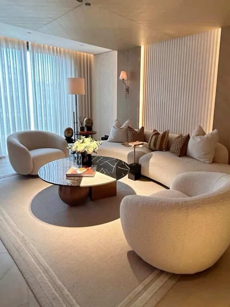 Modern living room with curved chairs, cozy sofa, round coffee table, flowers, and soft lighting.