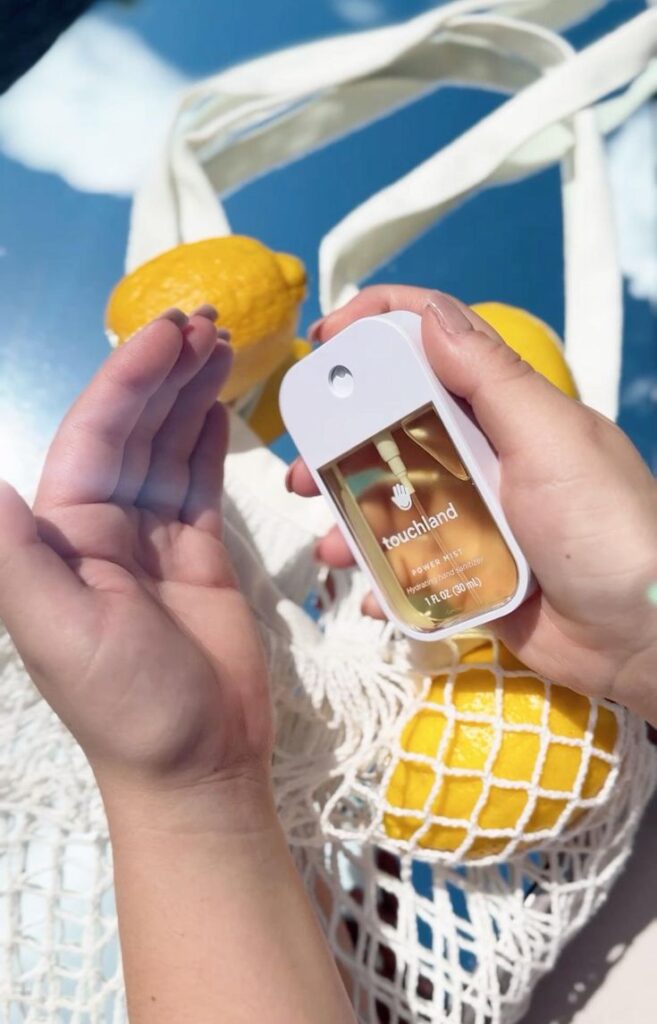 Hand holding Touchland sanitizer with lemons in a net bag on a sunny day.