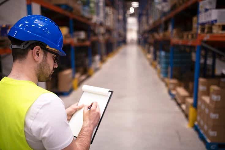 Warehouse worker in safety gear taking inventory notes in a large storage aisle.