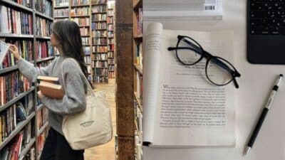 Person in a library browsing books, with open book, glasses, pen, and tablet on a desk.