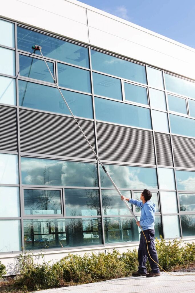 Person cleaning large modern office building windows with a long pole under a clear blue sky.