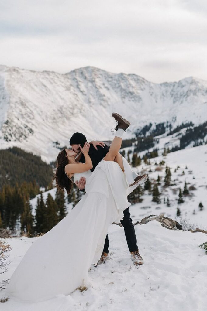 Couple embracing in snowy mountain landscape, woman lifted in a white dress, surrounded by scenic winter views.