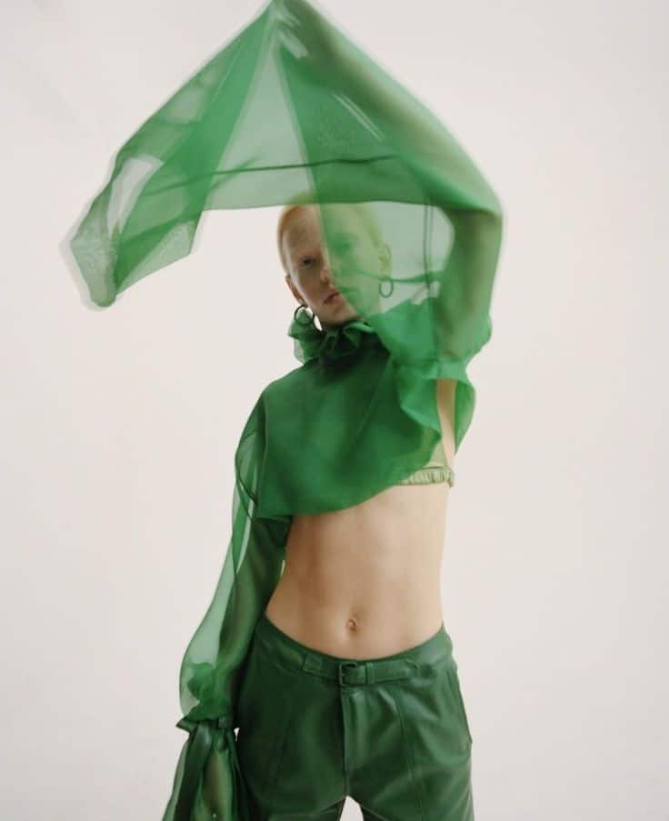 Person in flowing green outfit posing with a sheer fabric, modern fashion style, minimalistic background.