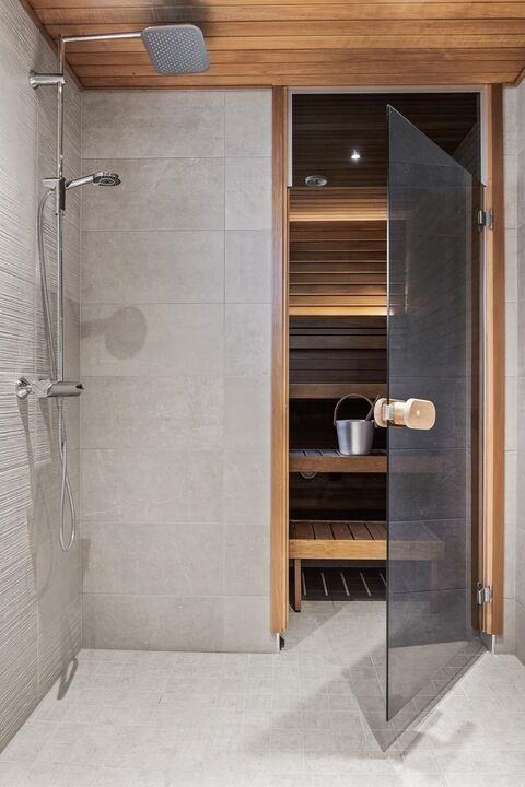 Modern bathroom with open glass door revealing a wooden sauna interior, featuring a shower and bucket.