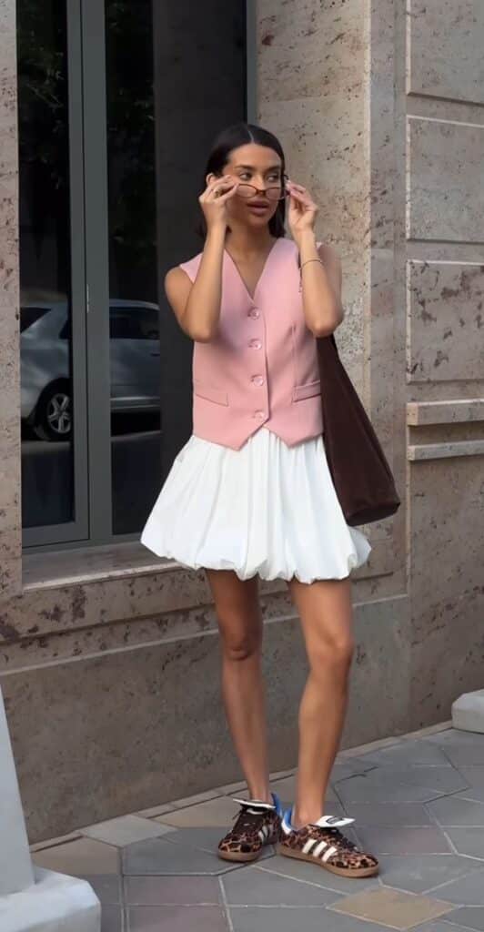 Trendy woman in pink vest, white skirt, and leopard sneakers, adjusts sunglasses while standing outdoors against stone wall.