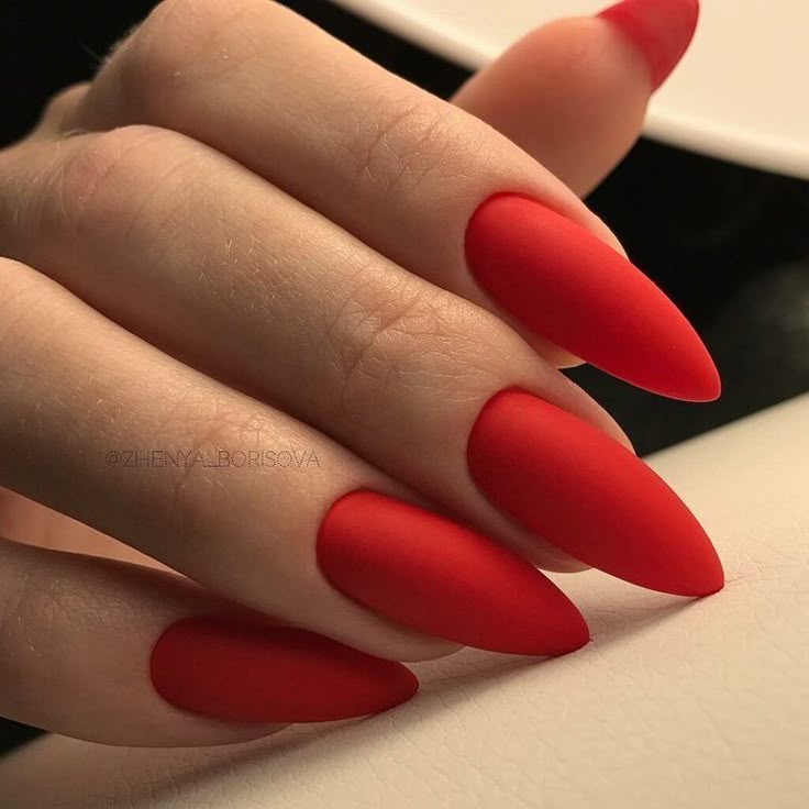 Close-up of hand with vibrant red, matte stiletto nails against a neutral background.