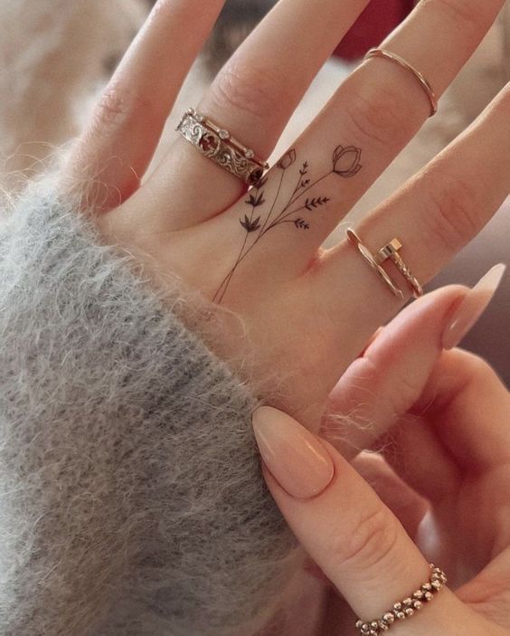 Close-up of a hand with floral tattoo and rings, wearing fuzzy grey sweater. Trendy minimalistic finger tattoo art.