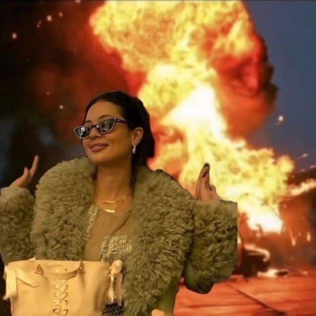 Woman with sunglasses and fur coat smiling confidently in front of a fiery explosion background.