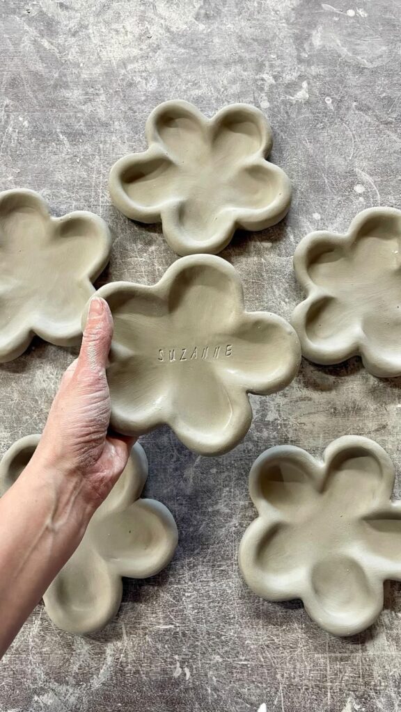 aa28d7795a3d89e72afbd439586fdbc2 Hand holding a clay flower-shaped dish labeled Suzanne among similar dishes on a textured background. | Sky Rye Design Hand holding a clay flower-shaped dish labeled Suzanne among similar dishes on a textured background.