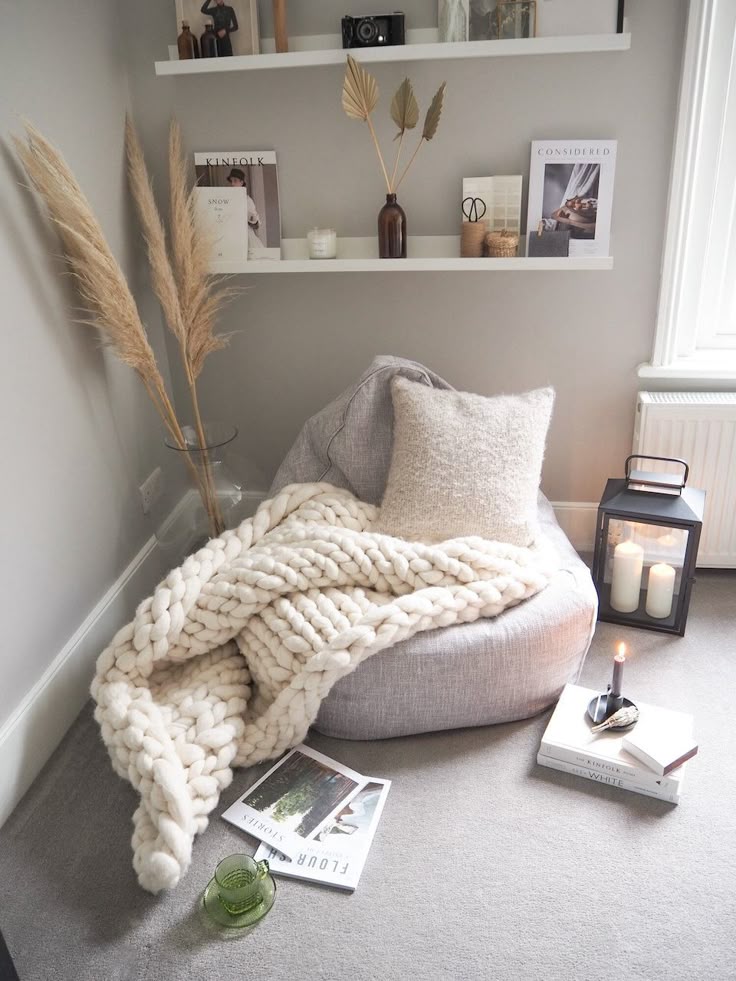 Cozy reading nook with a soft beanbag, chunky knit blanket, candles, and decorative shelves in a neutral-toned room.