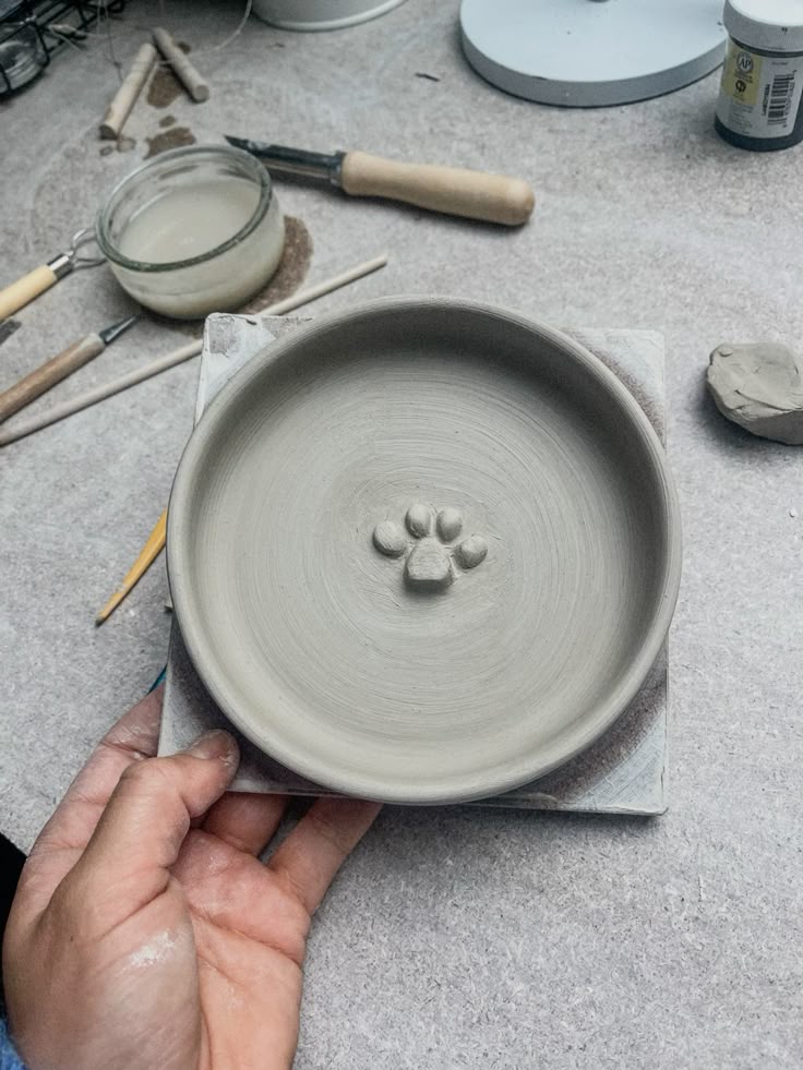 Hand holding a handcrafted clay bowl with a paw print design, surrounded by pottery tools on a workshop table.