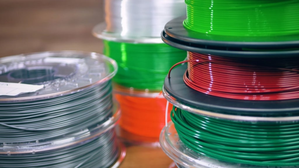 Colorful spools of 3D printer filament neatly stacked in a workshop, showcasing red, green, and gray plastic materials.