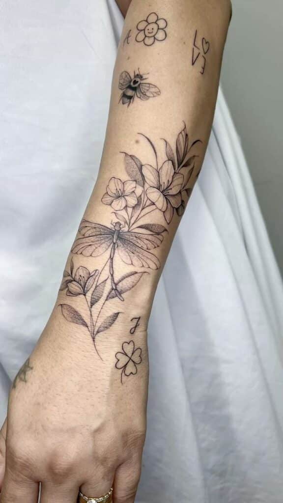 Forearm tattoos with dragonfly, flowers, bee, and heart design; delicate black ink art on skin.