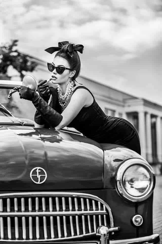 Vintage-inspired woman applying lipstick on classic car, embodying timeless elegance and retro fashion style.