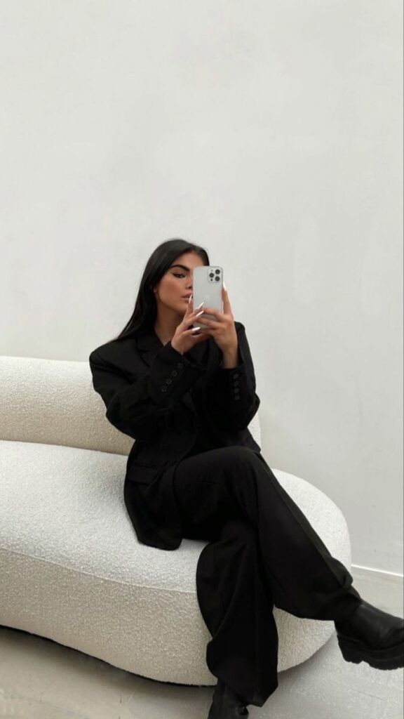Woman in black suit taking a mirror selfie on a white couch in minimalist setting.
