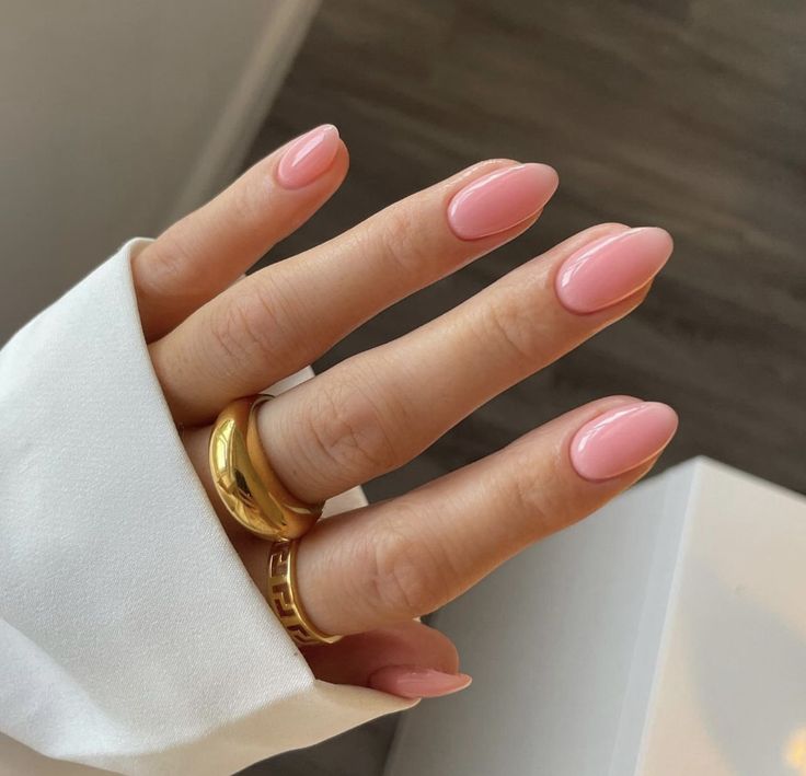 Pink almond-shaped nails with gold rings on hand against a white shirt background.