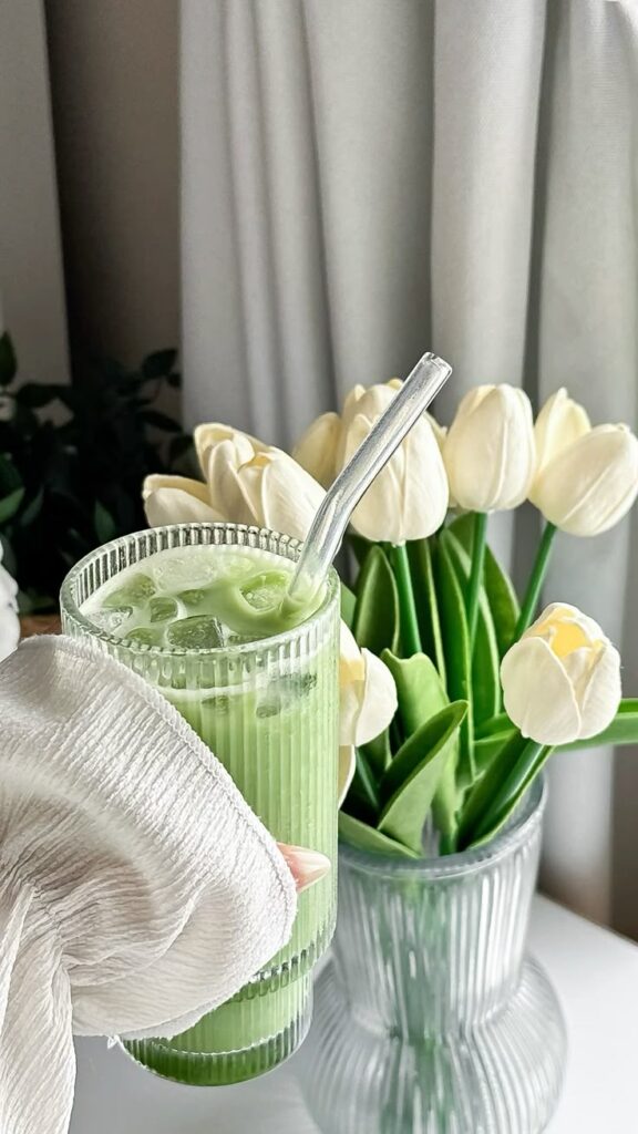 Green iced matcha latte in a ribbed glass with a straw, next to a vase of white tulips on a light background.