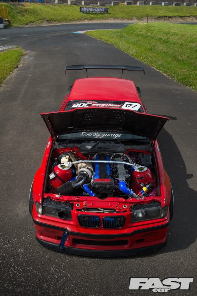 Red BMW drift car on track with hood open, showcasing modified engine. Racing decals on windshield, Fast Car logo.