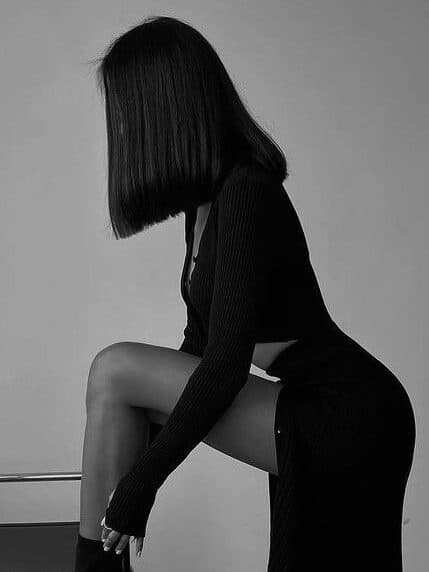 Woman in elegant black dress poses on a chair, showcasing modern fashion style. Black and white photography.
