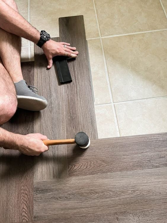 Man installing laminate flooring with a mallet and tapping block, over tiles.