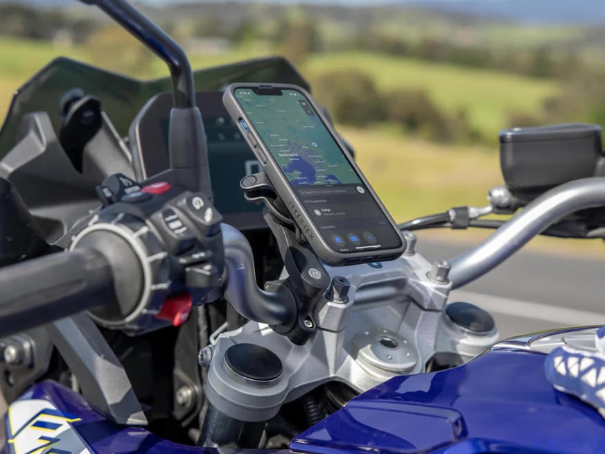 Smartphone mounted on motorcycle with GPS navigation displayed, scenic countryside visible in the background.