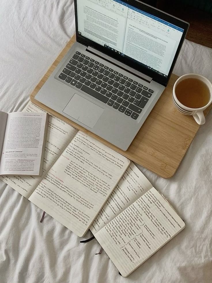 Laptop with documents, open books, and notes on a bed, accompanied by a cup of tea. Cozy study atmosphere.
