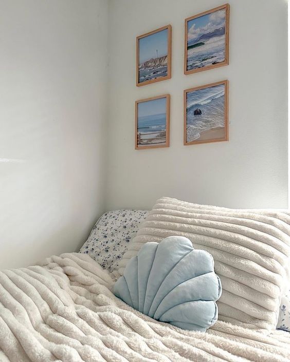 Cozy bedroom corner with soft pillows, fluffy blanket, and seascape photos on white walls. Inviting and serene decor.