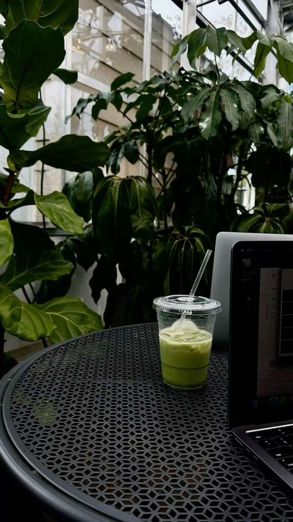 Laptop and iced green drink on a black metal table surrounded by lush green plants in a bright indoor setting.