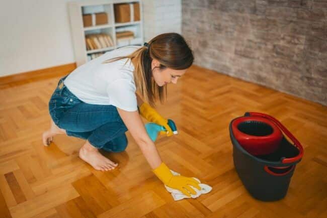 Woman cleaning wooden floor with cloth and spray bottle, using yellow gloves next to a red mop bucket.