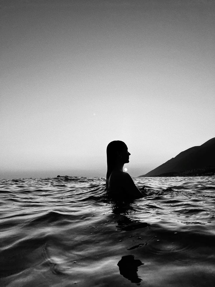 Silhouette of person in the ocean during sunset, with mountains in the background. Peaceful and serene black and white scene.