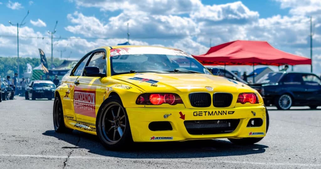 Yellow racing car with decals on display under a blue sky, parked at an outdoor auto show with tents and people.