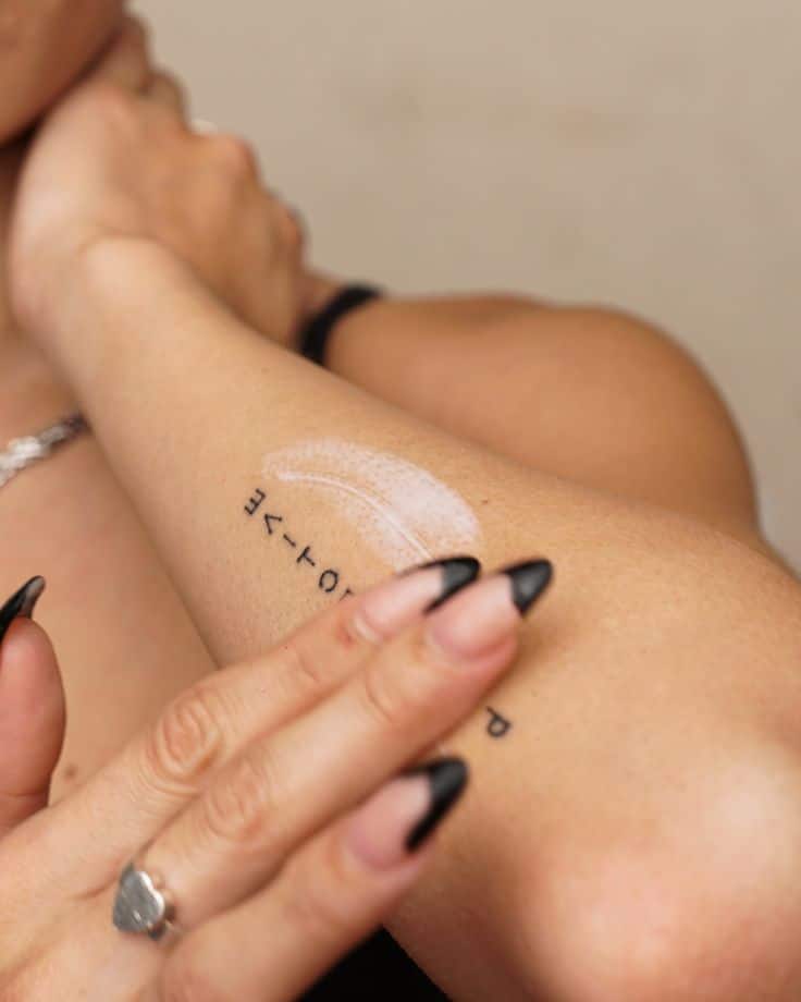 Person applying lotion on arm with tattoo and long nails.