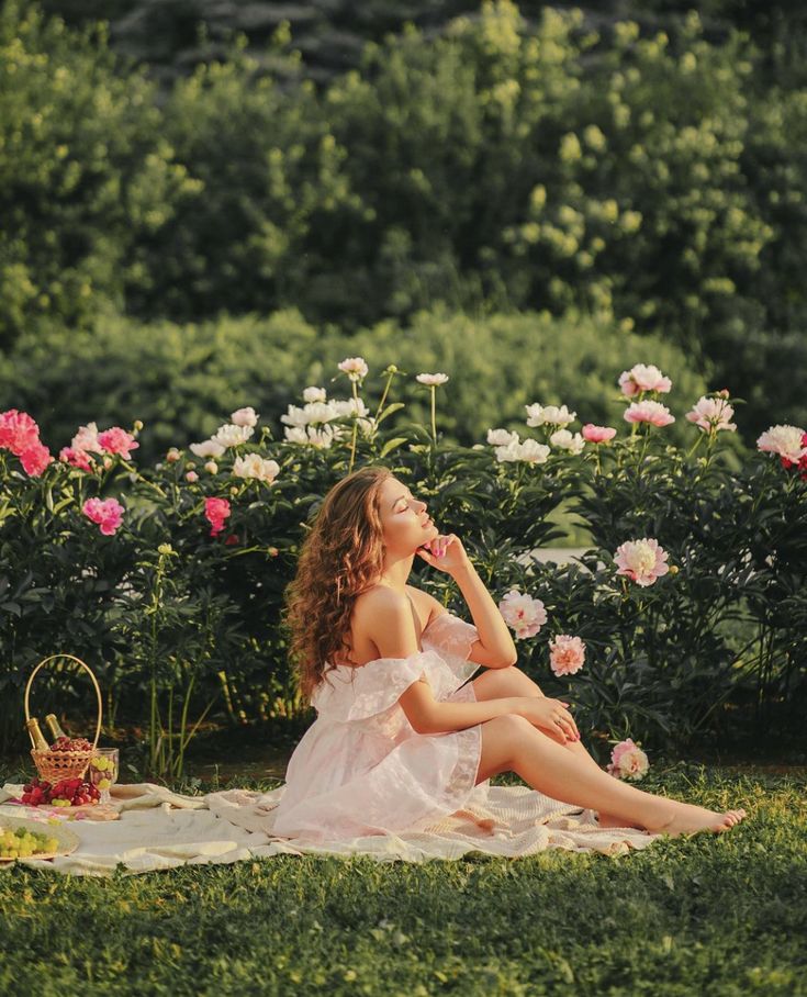 Woman in white dress enjoying a sunny picnic surrounded by colorful peonies in a lush garden.