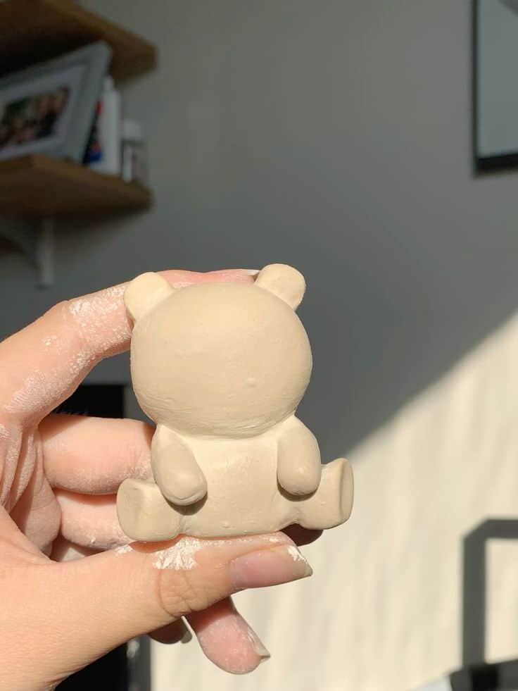 Hand holding a small, incomplete clay bear sculpture against a sunlit wall background.