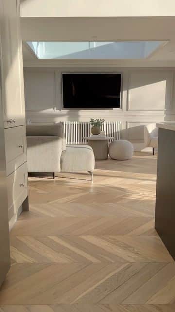 Modern living room with natural light, stylish furniture, wooden herringbone floor, and flat-screen TV on the wall.