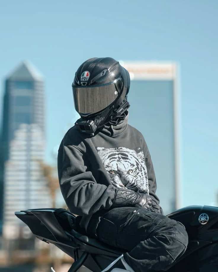 Motorcyclist in a sleek helmet sits on a bike against a cityscape backdrop, wearing a tiger-print hoodie.