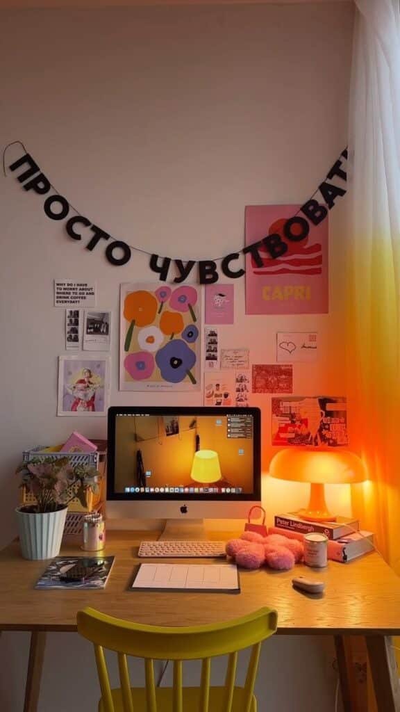 Cozy workspace with computer, warm lighting, posters, and decor on walls, featuring a yellow chair and desk accessories.