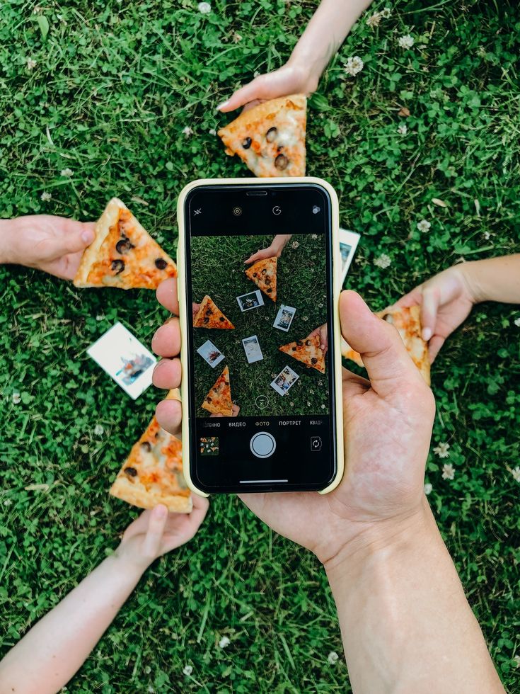 d49b03e0-7d13-4d82-98b1-703a1c94e2c4 Group sharing pizza slices outdoors while being photographed with a smartphone, creating a fun and social scene. | Sky Rye Design Group sharing pizza slices outdoors while being photographed with a smartphone, creating a fun and social scene.