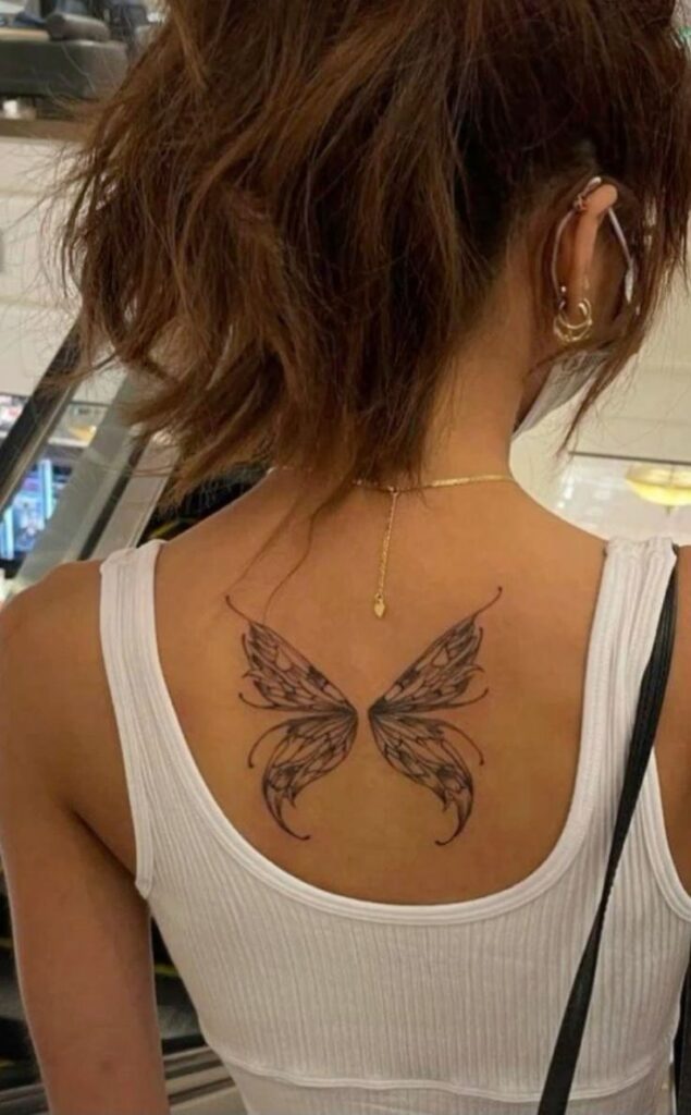 Butterfly tattoo on woman's upper back in a white tank top, showcasing intricate wing design and ear piercings.