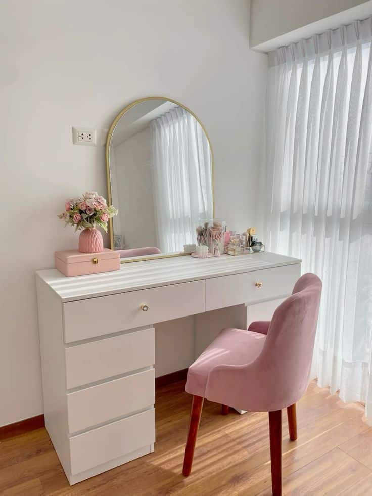 Chic vanity setup with white desk, pink chair, and gold mirror, adorned with flowers and makeup, wooden floor background.